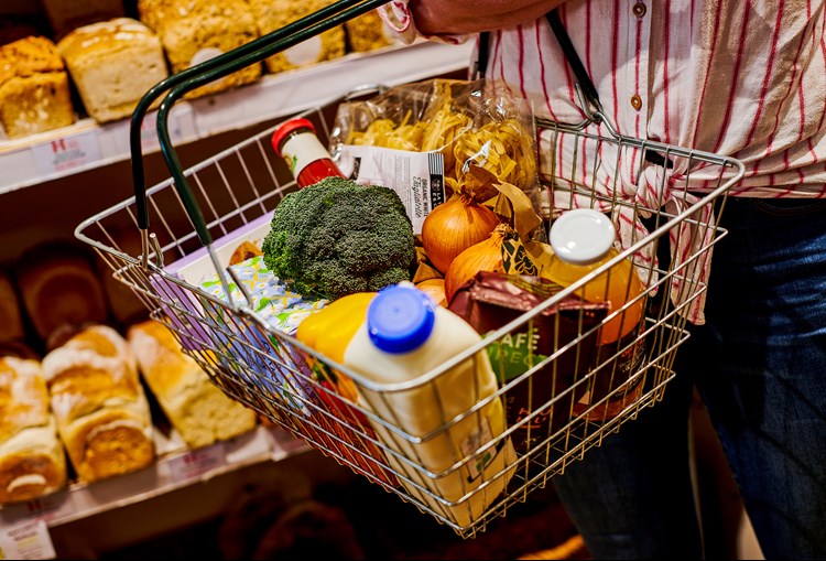 A person holding a shopping basket of food in front of a shelf of bread, with broccoli, milk, onions and other items in it